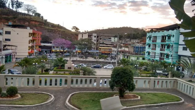 Praça de Inhapim MG ao entardecer, com jardim, bancos e o comércio colorido do centro ao fundo