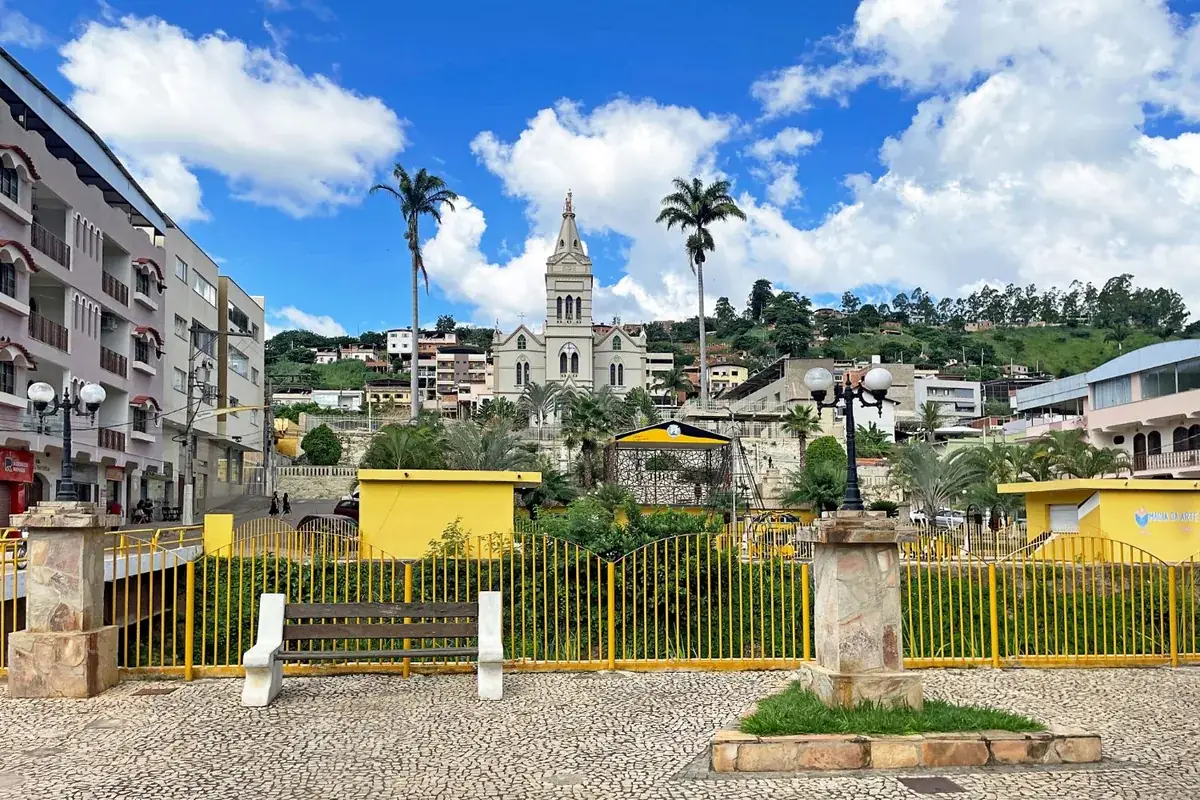 Praça da Matriz de Inhapim MG com gradil amarelo, bancos e palmeiras, e a torre da igreja matriz ao fundo